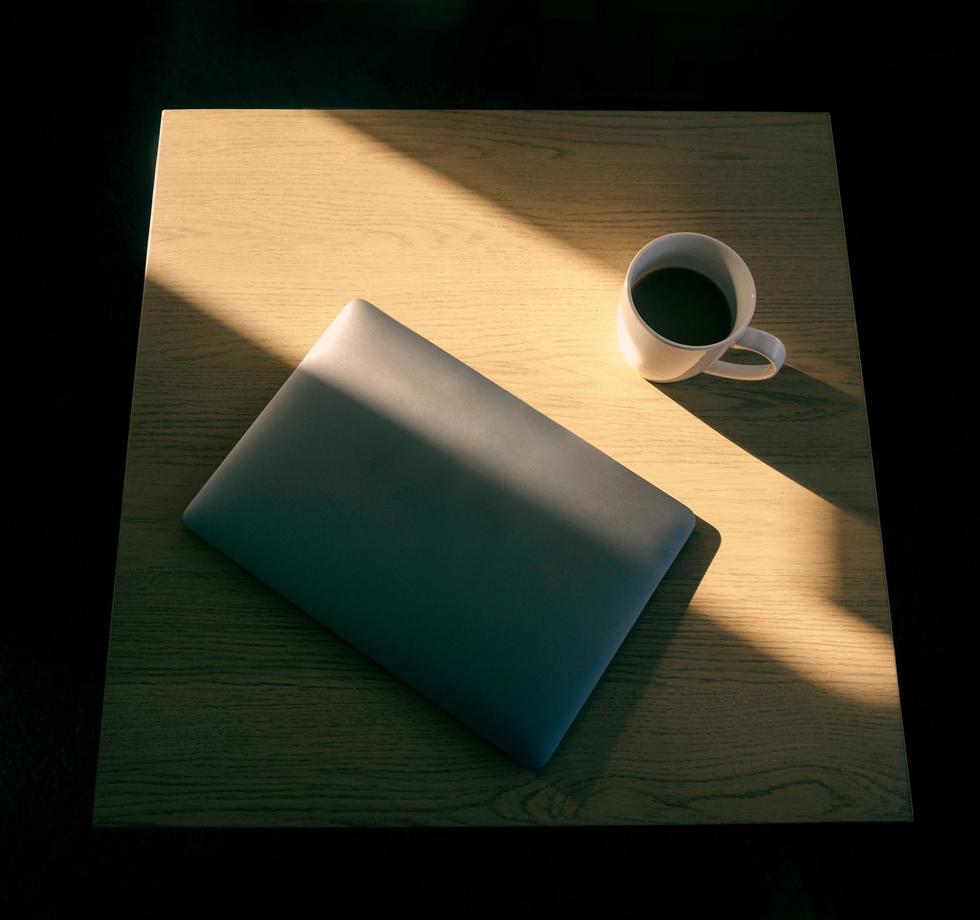 Un bureau minimaliste avec un ordinateur portable et une tasse de café sur une table en bois, jeu de lumière et d'ombre.