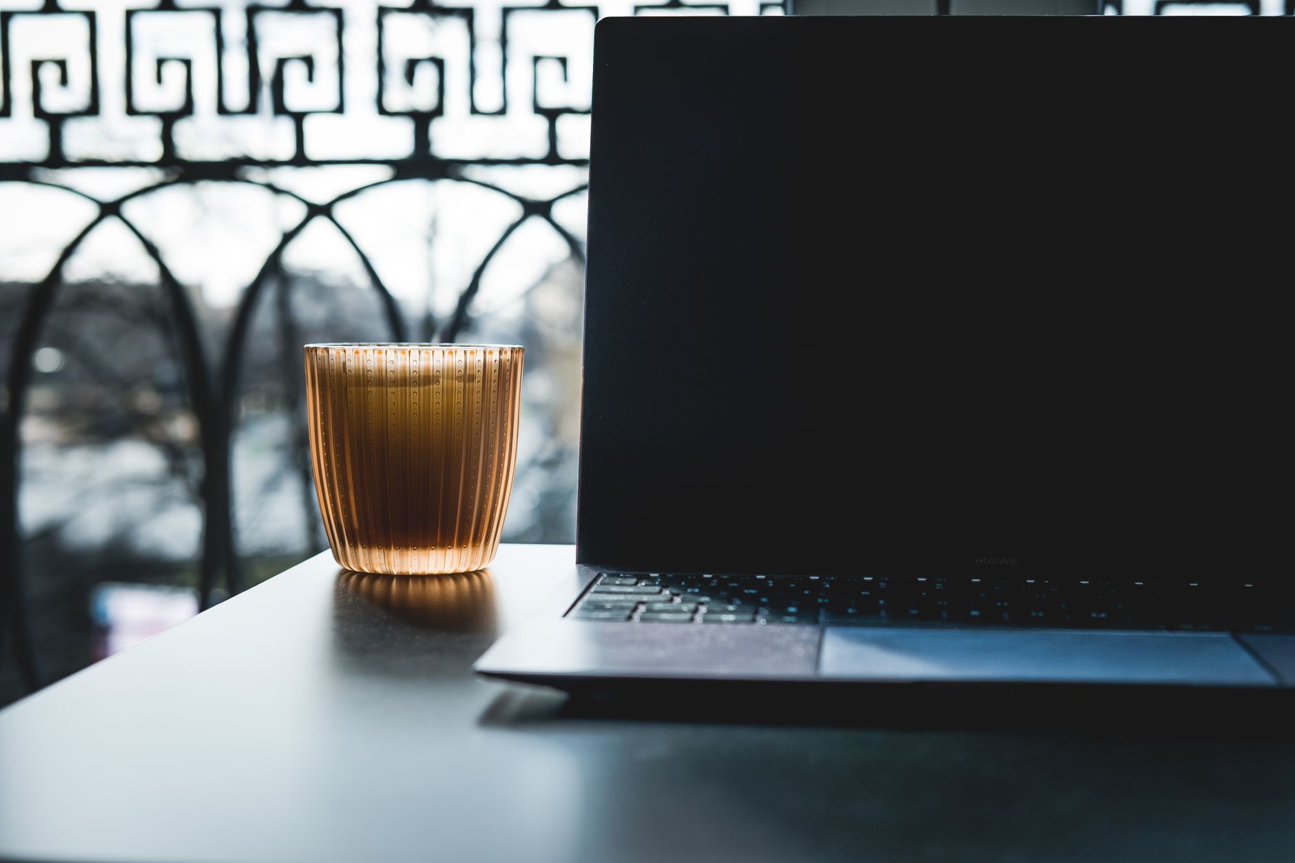 Un mini-ordinateur portable sur une table noire avec un verre de boisson, contre une balustrade de balcon ensoleillée.